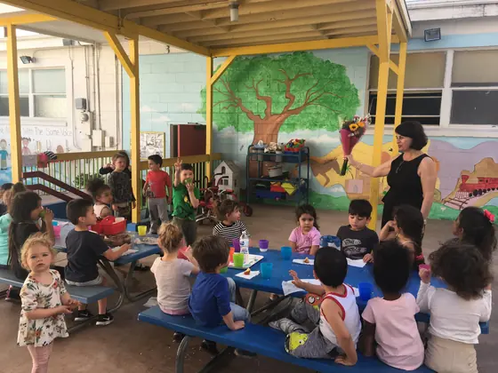 Children at snack time under the covered patio while a teacher holds flowers