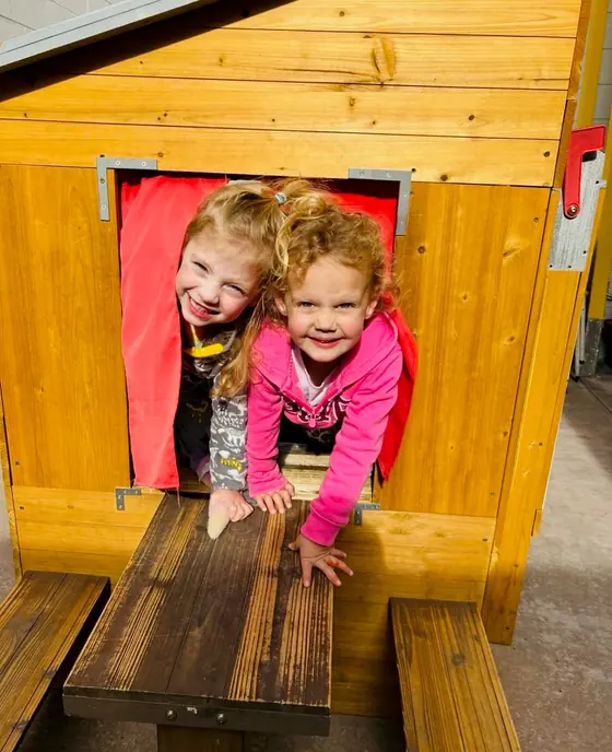 Two children smiling through the window of a wooden playhouse